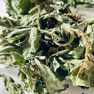 May include: Close-up of dried green leaves and stems, possibly herbs or botanicals. The leaves have a textured appearance with some curling. Small, dried seed heads are also visible, suggesting a natural product.