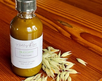 May include: A glass tincture bottle with a black dropper top, filled with a golden liquid. The label reads "Milky Oats Tincture" and "Fresh Milky Oat Tops, 60ml / 2 fl oz." Oat stalks are arranged beside the bottle on a wooden surface.