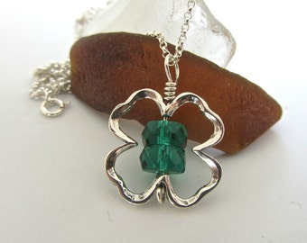 Puede incluir: Collar con colgante de trébol plateado con un centro de cuentas verde esmeralda facetadas. El colgante cuelga de una delicada cadena plateada. El collar se muestra sobre un fondo blanco con una piedra marrón y transparente.