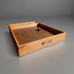 May include: Wooden dice tray with a brown felt interior and the text "Tavern Table". A single multi-sided die rests inside the tray. The tray is made of light-colored wood.