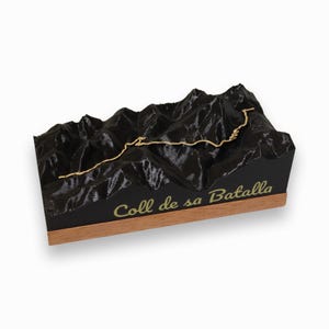 May include: A black 3D printed model of a mountain range with a gold-coloured road winding through it. The base of the model is made of wood and has the text "Coll de sa Batalla" inscribed on it.