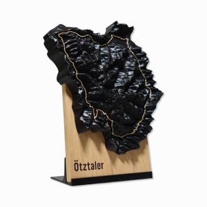 Può includere: Una mappa in legno 3D nera di una catena montuosa con una linea dorata che traccia un percorso. La mappa è montata su una base in legno con un supporto nero e il testo "Ötztaler" stampato sulla base.