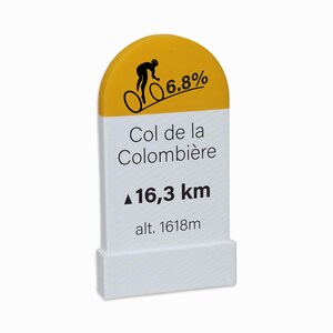 Könnte beinhalten: Ein weiß-gelbes, bogenförmiges Schild mit einer schwarzen Grafik eines Radfahrers, dem Text 6.8%, Col de la Colombière, 16,3 km und alt. 1618m. Das Schild hat einen rechteckigen Sockel und zeigt Informationen über einen Gebirgspass für Radfahrer.