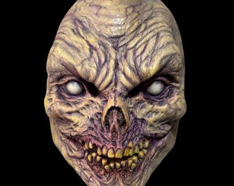 May include: A close-up of a detailed, grotesque, and realistic-looking monster mask. The mask is made of a textured material and has a pale yellow and purple color scheme. The mask has a wide, toothy grin and large, white eyes.