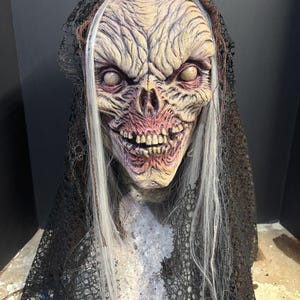 May include: A full-head mask featuring a weathered, wrinkled, pale-toned creature face with deep-set eyes, exposed teeth, and long, straight white hair. The mask is draped in a tattered, black, open-weave lace fabric that covers the head and shoulders.
