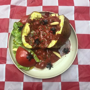 May include: A white plate with a red and brown baked potato, lettuce, tomato, and several plastic flies and maggots. The plate is on a red and white checked tablecloth.