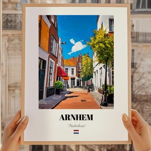Op de afbeelding: Een ingelijste print met een levendige straatscène in Arnhem, Nederland, onder een helderblauwe lucht. Het kunstwerk toont kleurrijke gebouwen, een rode luifel en een straat met bomen. Het woord "ARNHEM" wordt vetgedrukt weergegeven, met "Nederland" eronder.