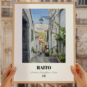 Può includere: Una stampa incorniciata di un dipinto raffigurante una stretta via italiana. L'opera presenta edifici bianchi, un cielo blu e la parola "RAITO". Il testo "Costiera Amalfitana, Italia" è anche visibile.