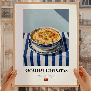 May include: A framed print featuring a still life of Bacalhau com Natas, a traditional Portuguese dish. The dish is in a blue and white bowl on a striped tablecloth. The print includes the text "BACALHAU COM NATAS" and "Made in Portugal."