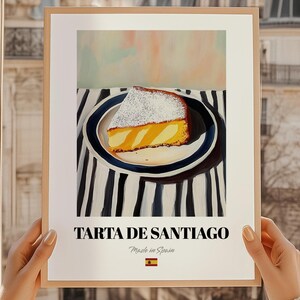 以下が含まれることがあります： スペインのアーモンドケーキ、タルタ・デ・サンティアゴの一切れが、青い縁の皿にのって描かれたフレームプリント。粉砂糖がまぶされています。「Tarta de Santiago」と「Made in Spain」の文字が入っています。