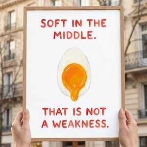 May include: A framed print with the text "SOFT IN THE MIDDLE. THAT IS NOT A WEAKNESS." The image shows a watercolour illustration of a soft-boiled egg with a runny yolk. The text and egg are in red and orange.