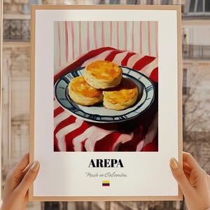 May include: A framed print featuring a still life of arepas, a traditional Colombian dish. The arepas are golden brown and stacked on a blue and white plate, set against a red and white striped background. The word "AREPA" is printed below the image.