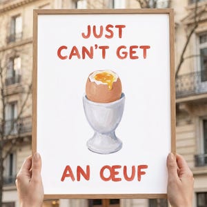 Könnte beinhalten: Ein gerahmter Druck mit den Worten "JUST CAN'T GET AN OEUF" in roten Buchstaben. Das Bild zeigt ein weich gekochtes Ei in einem weißen Eierbecher. Das Ei hat eine braune Schale und ein hellgelbes Eigelb.