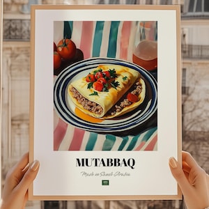 Op de afbeelding: Een ingelijste print met een culinaire illustratie van Mutabbaq, een hartige gevulde pannenkoek. Het gerecht wordt gepresenteerd op een blauw-wit gestreepte bord, gegarneerd met tomaten en peterselie. De print bevat de tekst "MUTABBAQ" en "Made in Saudi Arabia".