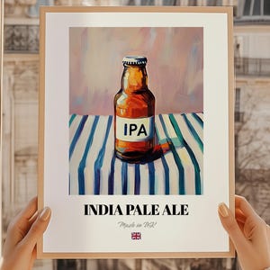 May include: Framed print featuring an IPA beer bottle on a blue and white striped cloth. The label reads "IPA". Below the image are the words "INDIA PALE ALE" and "Made in UK" with a British flag, indicating its origin.
