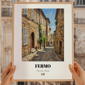 Puede incluir: Una impresión enmarcada de una escena callejera italiana en Fermo, Marche, Italia. La obra de arte representa una calle estrecha adoquinada bordeada de edificios antiguos. La impresión está sostenida por una persona con uñas cuidadas. La palabra "FERMO" está impresa en negrita, en negro.