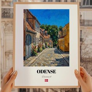 Könnte beinhalten: Ein gerahmter Druck einer europäischen Straßenszene. Das Kunstwerk zeigt eine Kopfsteinpflasterstraße mit farbenfrohen Gebäuden unter einem strahlend blauen Himmel. Das Wort "ODENSE" ist unten aufgedruckt, zusammen mit dem Wort "Dänemark" und der dänischen Flagge.