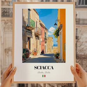 May include: A framed print of a colorful street scene in Sciacca, Sicily, Italy. The artwork depicts buildings in shades of orange, pink, and beige, with balconies and potted plants. The title "Sciacca" is printed in black, with the Italian flag below.