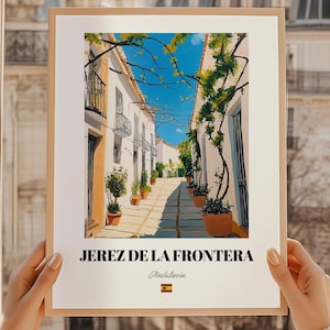May include: A framed print featuring a sunlit street scene in Jerez de la Frontera, Spain. The artwork depicts white buildings with balconies, potted plants, and a cobblestone path. The print includes the text "JEREZ DE LA FRONTERA" and "Andalucia".