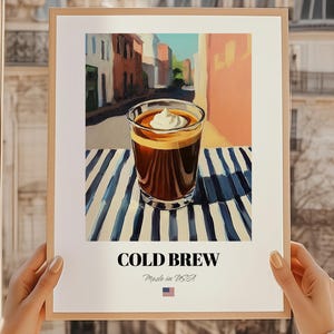 Op de afbeelding: Een ingelijste kunstdruk met een glas cold brew koffie met slagroom, tegen een straatbeeld. De print heeft een witte rand en de woorden "COLD BREW" staan onderaan gedrukt, samen met "Made in USA" en de Amerikaanse vlag.