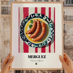 Könnte beinhalten: Gerahmter Kunstdruck mit drei gekochten Würstchen auf einem dekorativen Teller. Der Teller befindet sich vor einem Hintergrund aus roten und weißen vertikalen Streifen. Das Wort "MERGUEZ" ist unten zusammen mit "Made in Algeria" aufgedruckt.