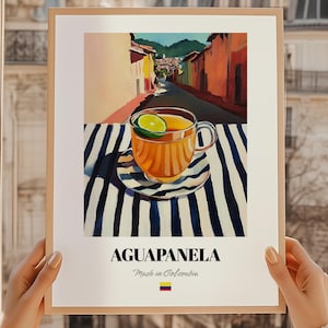 Puede incluir: Una impresión artística enmarcada que presenta una ilustración de una taza de aguapanela con una rodaja de lima, sobre una superficie a rayas. El fondo representa una colorida escena callejera. El texto en la parte inferior dice "AGUAPANELA" y "Made in Colombia".
