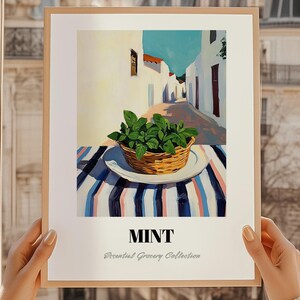May include: A framed print showing a basket of mint on a striped tablecloth, set against a sunlit alleyway with white buildings and a blue sky. The word "MINT" is displayed below the image, with "Essential Grocery Collection" underneath.