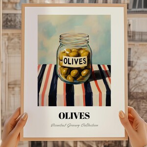 May include: A framed print depicting a jar of olives with a label reading "OLIVES." The jar is placed on a table with a striped pattern of red, white, and navy blue. The words "OLIVES" and "Essential Grocery Collection" are printed below the image.