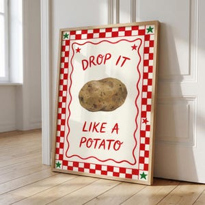 Puede incluir: Impresión artística enmarcada con una ilustración de patata y el texto "DROP IT LIKE A POTATO" en rojo. La obra de arte tiene un fondo blanco con un borde a cuadros rojo y blanco y acentos de estrella, en un marco de madera.