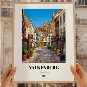 以下が含まれることがあります： オランダのファルケンブルクのストリートシーンを描いた額入りのプリント。カラフルな建物、ショップ、カフェが並ぶ石畳の通りが描かれています。「VALKENBURG Nederland」の文字が下部に表示され、オランダの国旗も添えられています。