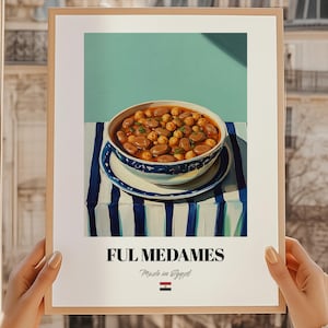 Peut inclure: Une affiche encadrée représentant un bol de ful medames, un plat égyptien traditionnel. Le bol est posé sur une surface à rayures bleues et blanches, sur fond turquoise. Le texte "FUL MEDAMES Made in Egypt" est affiché en dessous.