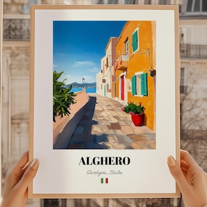 May include: A framed art print featuring a vibrant street scene in Alghero, Italy. The artwork depicts colorful buildings with orange, white, and teal accents, a cobblestone street, and a view of the sea under a bright blue sky. The print includes the text "ALGHERO".