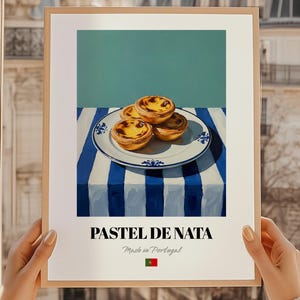 Puede incluir: Una impresión enmarcada con pastel de nata, una tarta de crema portuguesa, sobre un mantel a rayas azules y blancas. El plato tiene un diseño floral azul. El texto "PASTEL DE NATA" se muestra debajo de la imagen, con "Made in Portugal" y la bandera portuguesa.
