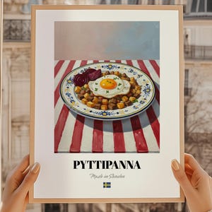 Op de afbeelding: Een ingelijste print van een gerecht met een gebakken ei, aardappelen en rode bieten. Het bord staat op een rood-wit gestreept tafelkleed. De print bevat het woord "PYTTIPANNA" en "Made in Sweden" eronder.