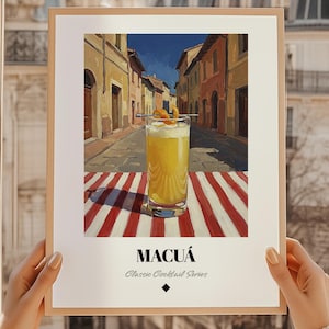 Puede incluir: Una impresión enmarcada de un cóctel, el Macuá, de la serie Classic Cocktail. La imagen muestra un vaso alto de cóctel amarillo con una guarnición, sobre una superficie a rayas rojas y blancas, con una escena de calle europea de fondo.
