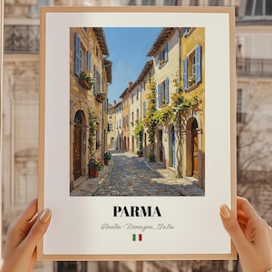 Puede incluir: Una impresión enmarcada de una escena de calle europea. La obra de arte representa una calle estrecha adoquinada bordeada de edificios en tonos amarillos y beige, con contraventanas azules y jardineras. La palabra "PARMA" está impresa en la parte inferior.
