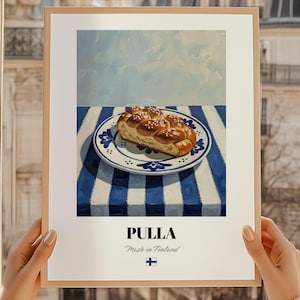 Op de afbeelding: Een ingelijste print met een stilleven van een plak pulla, een Fins zoet brood, op een blauw-wit gestreept tafelkleed. Het brood ligt op een decoratief bord. Het woord "PULLA" staat onderaan afgedrukt, met daaronder "Made in Finland".