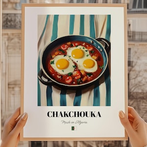 May include: A framed print featuring a vibrant illustration of shakshuka, a Middle Eastern and North African dish. The artwork depicts three sunny-side-up eggs in a rich tomato sauce, served in a blue pan. The print includes the word "CHAKCHOUKA" and "Made in Algeria".