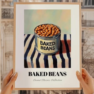 Op de afbeelding: Een ingelijste print met een blikje baked beans. Het blikje, met het opschrift "BAKED BEANS", is gevuld met bonen en staat op een gestreept oppervlak. De print maakt deel uit van de "Canned Classics Collection".