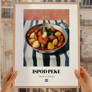 May include: A framed print featuring a vibrant illustration of a stew with potatoes, carrots, and meat in a silver pan. The print is set against a striped blue and white tablecloth. The text "ISPOD PEKE" is displayed below the image.