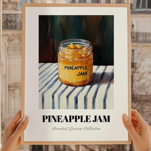 May include: A framed print featuring a jar of pineapple jam on a striped surface. The jar has a label that reads "PINEAPPLE JAM." Below the jar, the text "PINEAPPLE JAM" is displayed, with "Essential Grocery Collection" underneath. The print is held by a person.