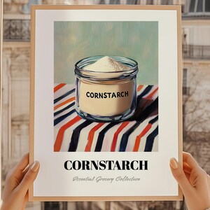 May include: A framed print showing a jar of cornflour on a striped surface. The clear jar contains white powder, with "CORNSTARCH" printed on it. The print is part of the "Essential Grocery Collection".