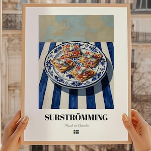 May include: A framed print featuring a still life of Swedish surströmming. The image shows a blue and white patterned plate with four open-faced sandwiches topped with fish, tomatoes, and onions. The print has the word "SURSTRÖMMING" and "Made in Sweden" printed on it.