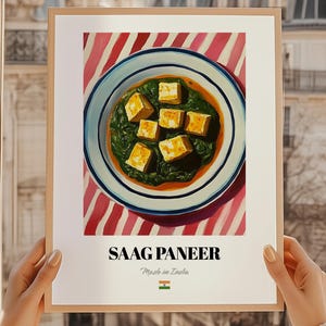Könnte beinhalten: Ein gerahmter Druck von Saag Paneer, einem beliebten indischen Gericht. Das Kunstwerk zeigt eine Schüssel des Gerichts mit Paneer-Käsewürfeln in einer grünen Spinatsoße, vor einem rot-weiß gestreiften Hintergrund. Der Text "SAAG PANEER" und "Made in India" stehen unten.