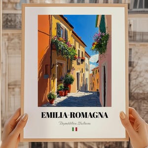 Può includere: Una stampa incorniciata raffigurante una vivace scena di strada dell'Emilia-Romagna, Italia. L'opera d'arte mostra edifici nei toni dell'arancione, giallo e rosa, con fioriere e piante in vaso. Il testo "EMILIA-ROMAGNA" è visualizzato in basso, insieme al testo italiano e alla bandiera italiana.