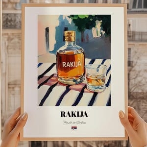 Könnte beinhalten: Gerahmter Kunstdruck mit einer Flasche und einem Glas Rakija. Die Flasche ist bernsteinfarben mit einem goldenen Verschluss, das Glas ist teilweise gefüllt. Das Wort "RAKIJA" steht am unteren Rand des Drucks.