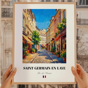 May include: A framed art print of a European street scene. The artwork depicts a cobblestone street lined with colorful buildings, shops, and trees under a bright blue sky. The print includes the text "SAINT-GERMAIN-EN-LAYE" and the French flag.
