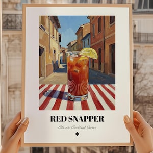 May include: A framed print of a Red Snapper cocktail from the Classic Cocktail Series. The image features a tall glass filled with a reddish-brown drink, garnished with a lemon slice, set against a backdrop of a European street scene. The drink sits on a red and white striped surface.