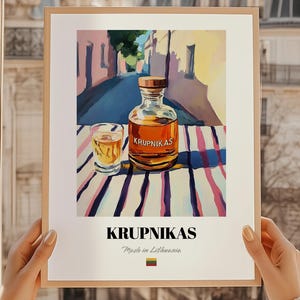 May include: A framed print featuring a still life of a bottle and a shot glass of amber liquid, labeled "Krupnikas." The background depicts a street scene. The print is held by a person.