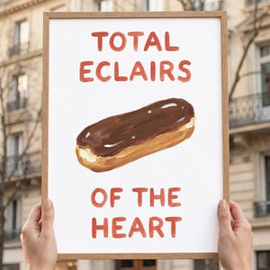 May include: A framed print with the words "TOTAL ECLAIRS OF THE HEART" in red lettering. The print features a watercolour painting of a chocolate éclair. The frame is a light brown colour.